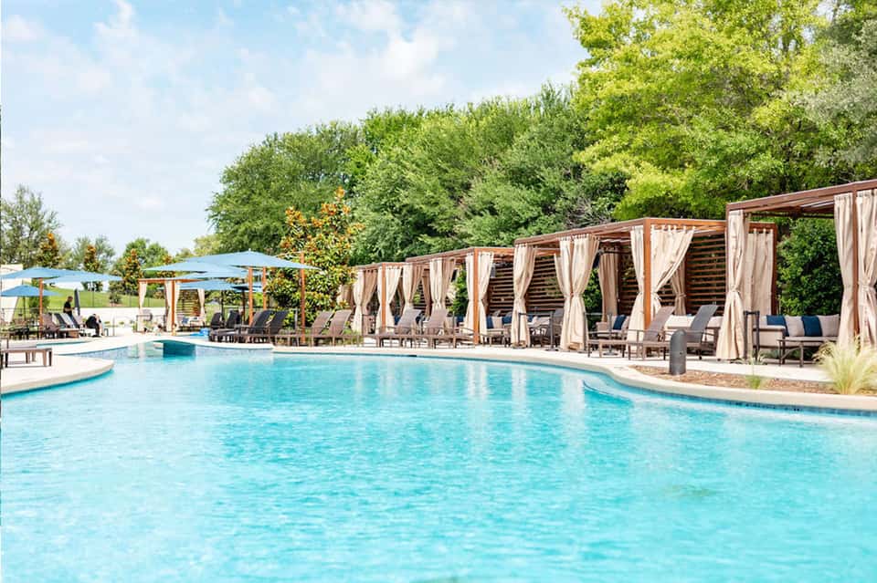 Resort pool with wooden pergolas draped in white curtains, lounge chairs, and lush green trees in background