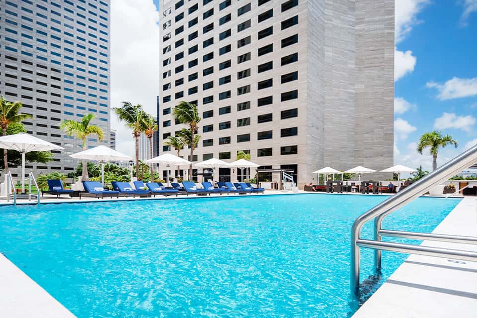 Resort pool with blue loungers, white umbrellas, and modern high-rise hotel buildings