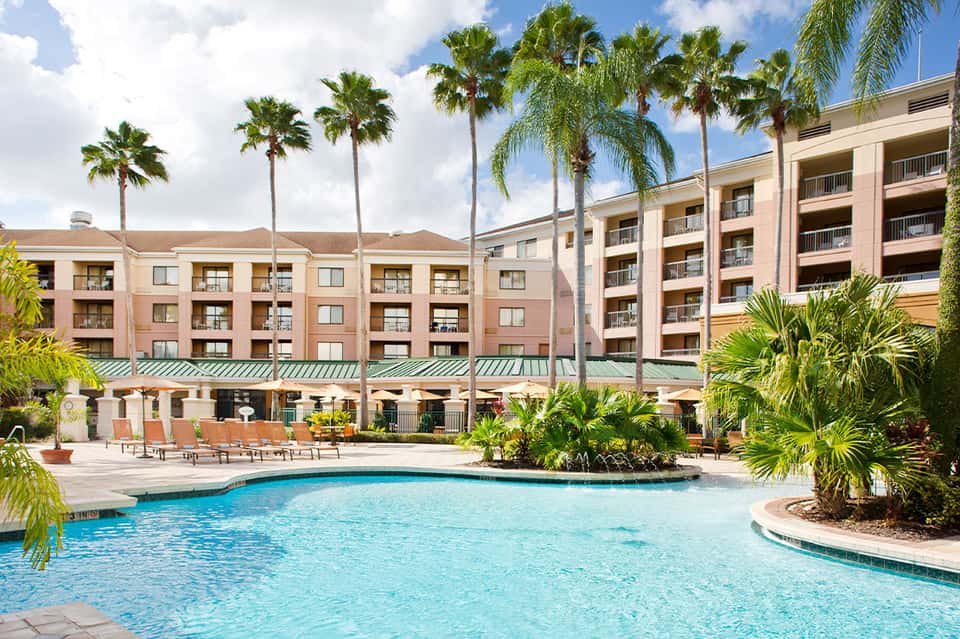 Resort courtyard with sparkling blue pool, palm trees, and multi-story pink hotel buildings with balconies