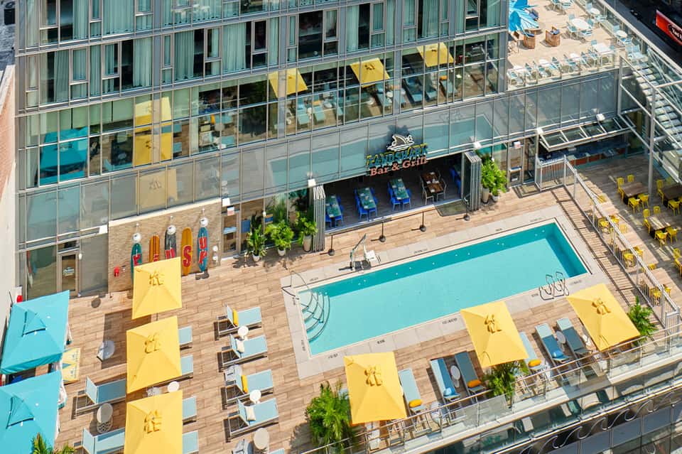 Rooftop pool deck with turquoise swimming pool, yellow umbrellas, lounge chairs, and high-rise building facade
