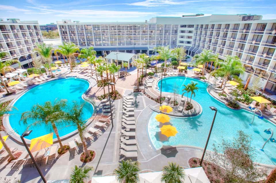 Aerial view of resort with two turquoise pools, palm trees, and modern apartment buildings surrounding a central courtyard