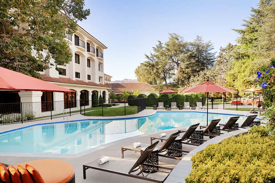 Curved outdoor pool with lounge chairs, red umbrellas, and Spanish-style hotel building