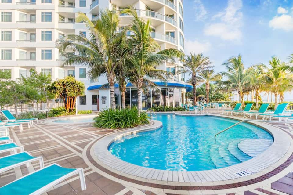 Resort pool with curved sections, lounge chairs, palm trees, and modern white building