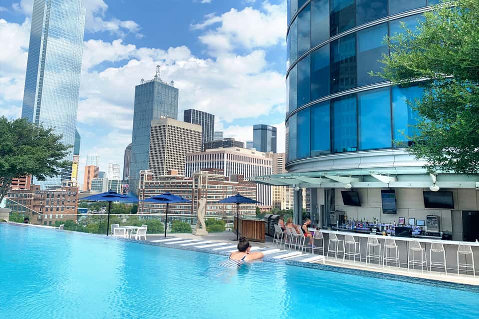 Rooftop infinity pool with city skyline views, guests swimming, blue umbrellas, and bar seating area