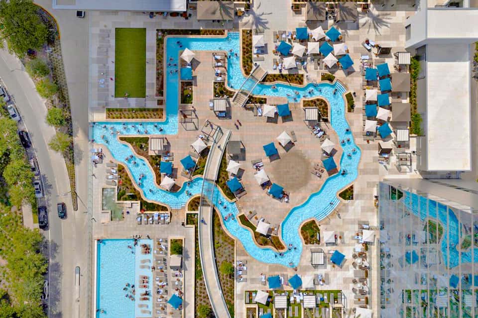 Aerial view of resort pool complex with blue water features, loungers with umbrellas, and surrounding buildings