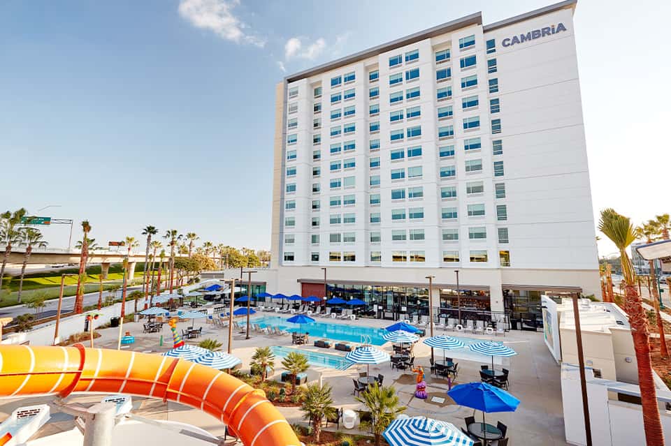 Modern hotel exterior with blue pool, orange slide, umbrellas, and palm trees surrounding white building