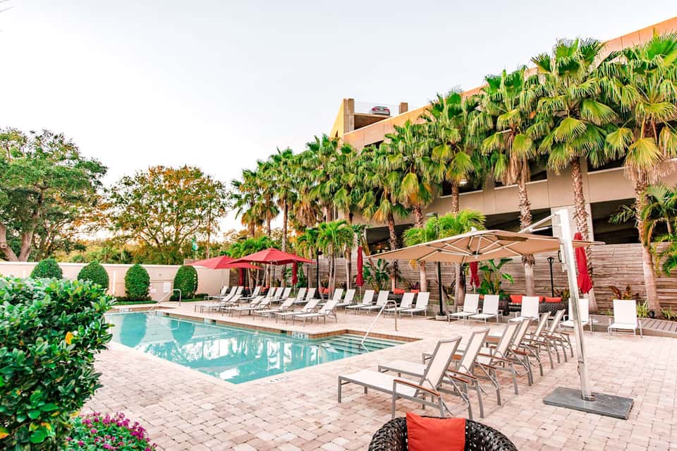Resort pool area with lounge chairs, red umbrellas, and palm trees surrounding a rectangular swimming pool