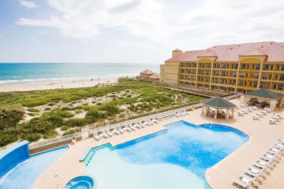 Beachfront resort pool with yellow building, sandy beach, ocean view, and lounge chairs