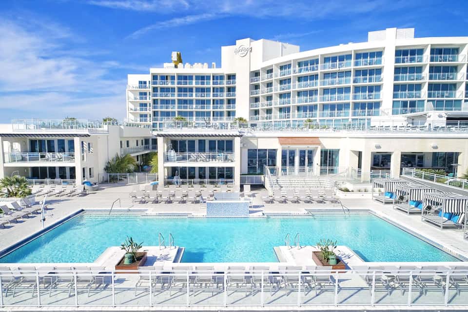 Beachfront hotel pool area with blue water, white loungers, and modern white building