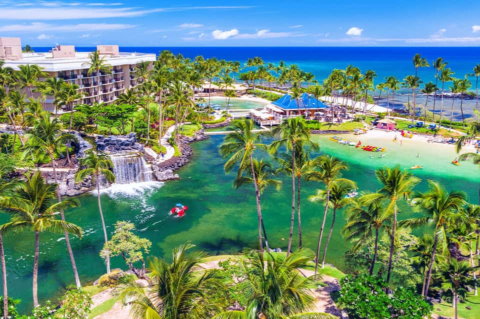Aerial view of beachfront resort with lagoon pool, palm trees, white sand beach, and turquoise ocean