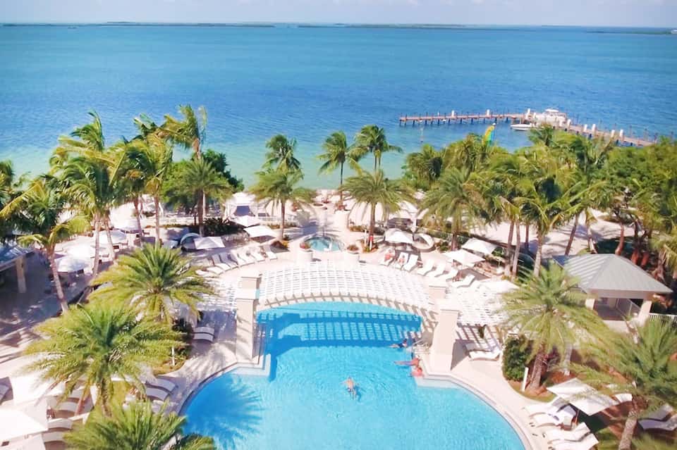 Aerial view of beachfront resort with curved pool, palm trees, and ocean pier