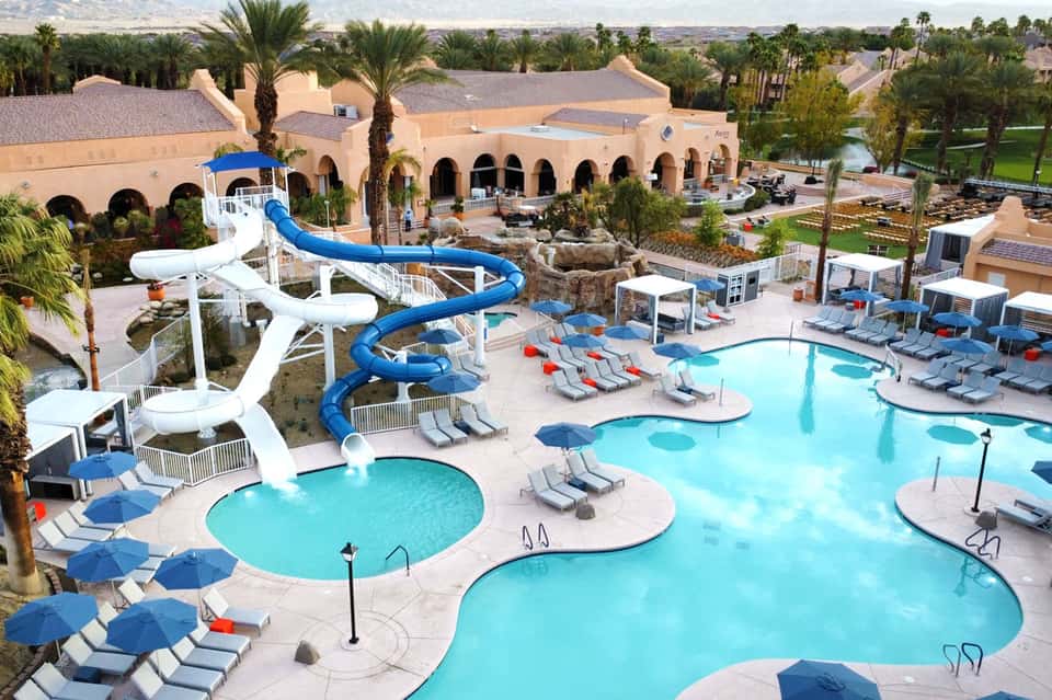 Aerial view of resort with multiple turquoise pools, water slides, loungers, and tan Moroccan-style buildings surrounded by palm trees
