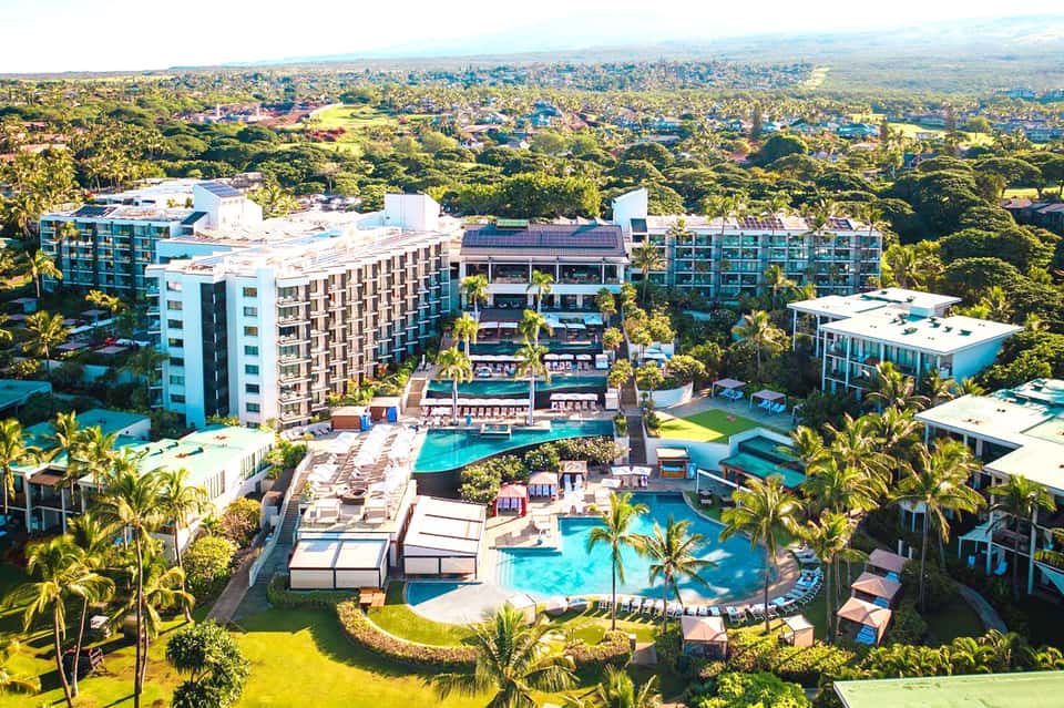 Aerial view of large resort complex with multiple pools, palm trees, and surrounding green landscape