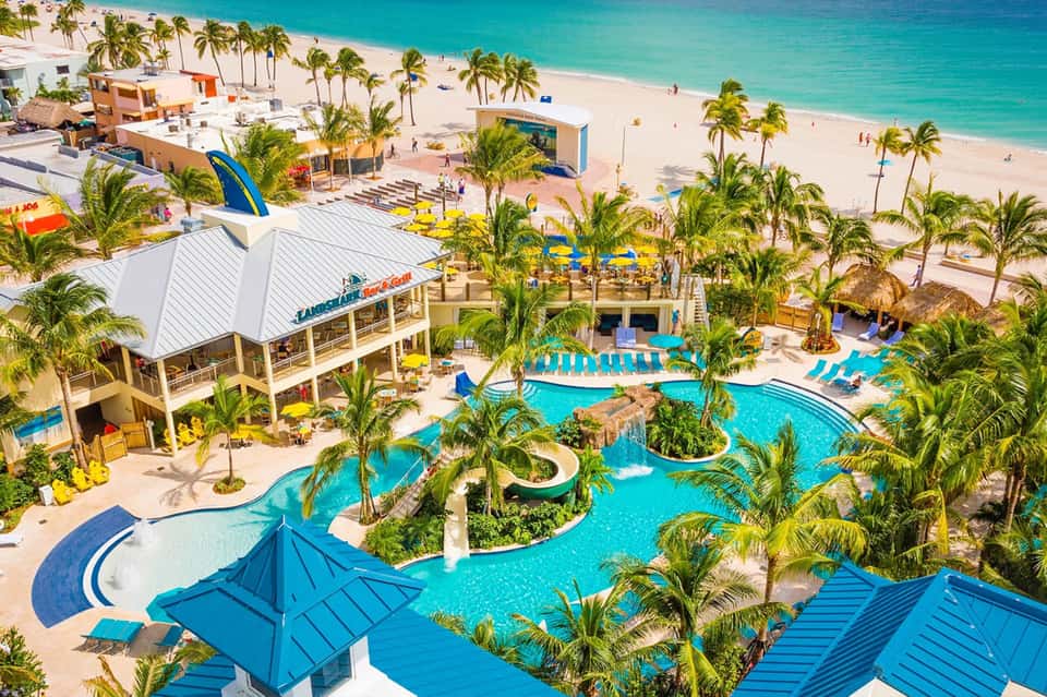 Aerial view of beachfront resort with turquoise pools, palm trees, water slides, and sandy beach with ocean beyond