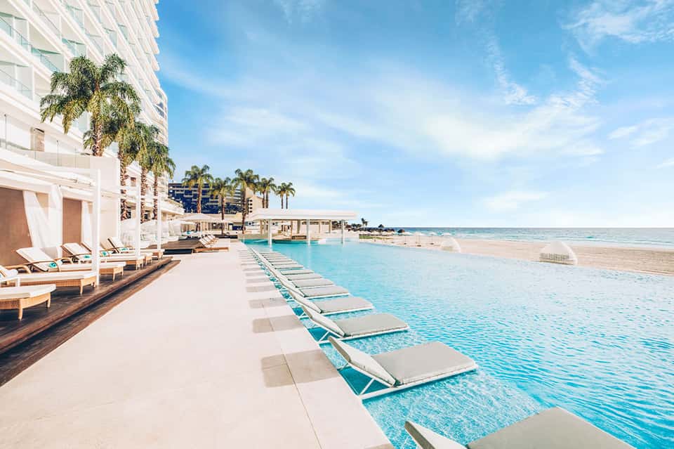 Beachfront resort with lounge chairs lining an infinity pool overlooking the ocean and palm trees