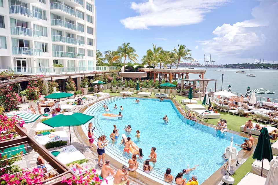 Resort pool with turquoise water, loungers, umbrellas, waterfront buildings, and boats in harbor