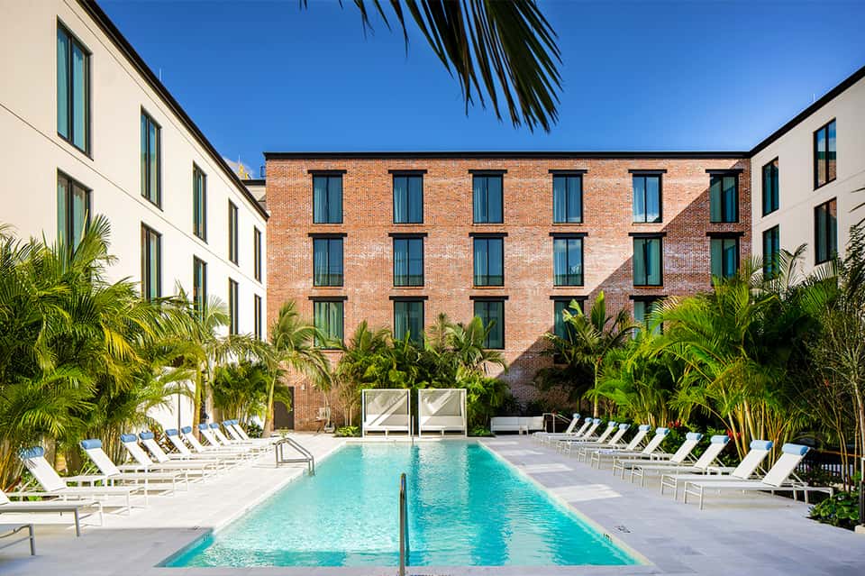 Resort courtyard pool surrounded by white lounge chairs, palm trees, and brick buildings