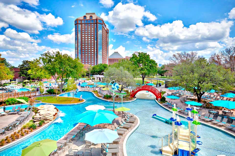 Large resort pool complex with blue water, red bridge, colorful umbrellas, and tall red brick hotel building