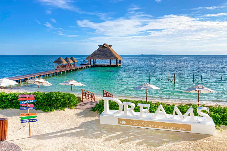 Pristine beach with DREAMS resort sign, overwater bungalows, dock, and white sand