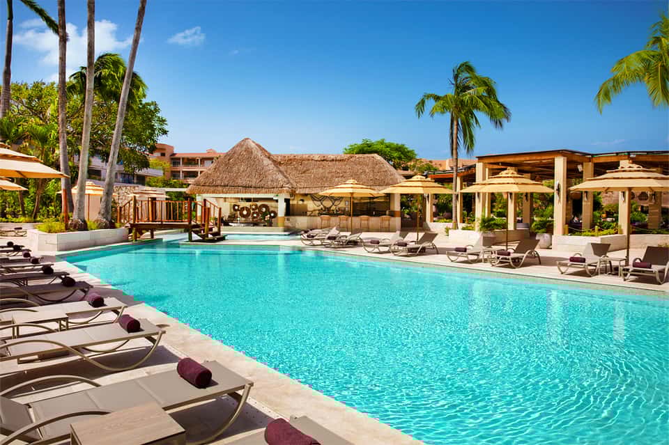 Luxury resort pool with turquoise water, palm trees, and thatched-roof structures