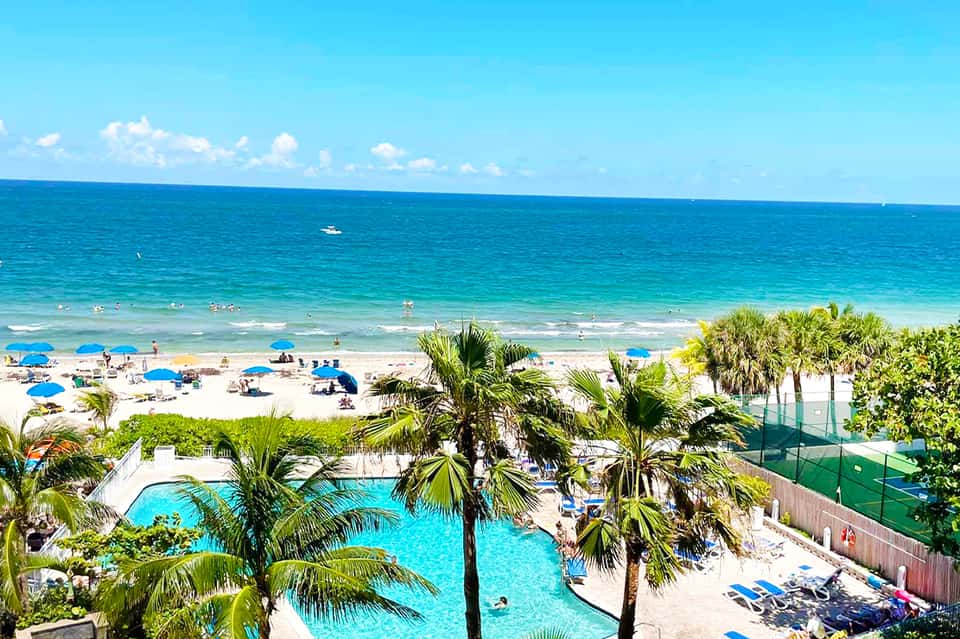 Beachfront resort pool with palm trees, sandy beach, and ocean view under blue sky