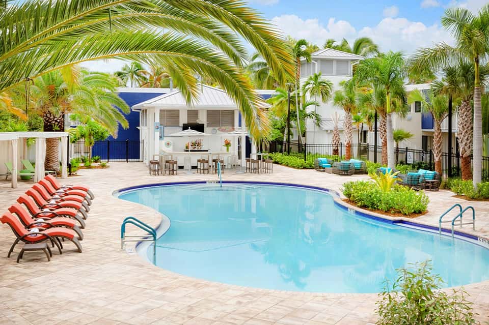 Large resort pool with curved shape, red lounge chairs, white buildings, and palm trees