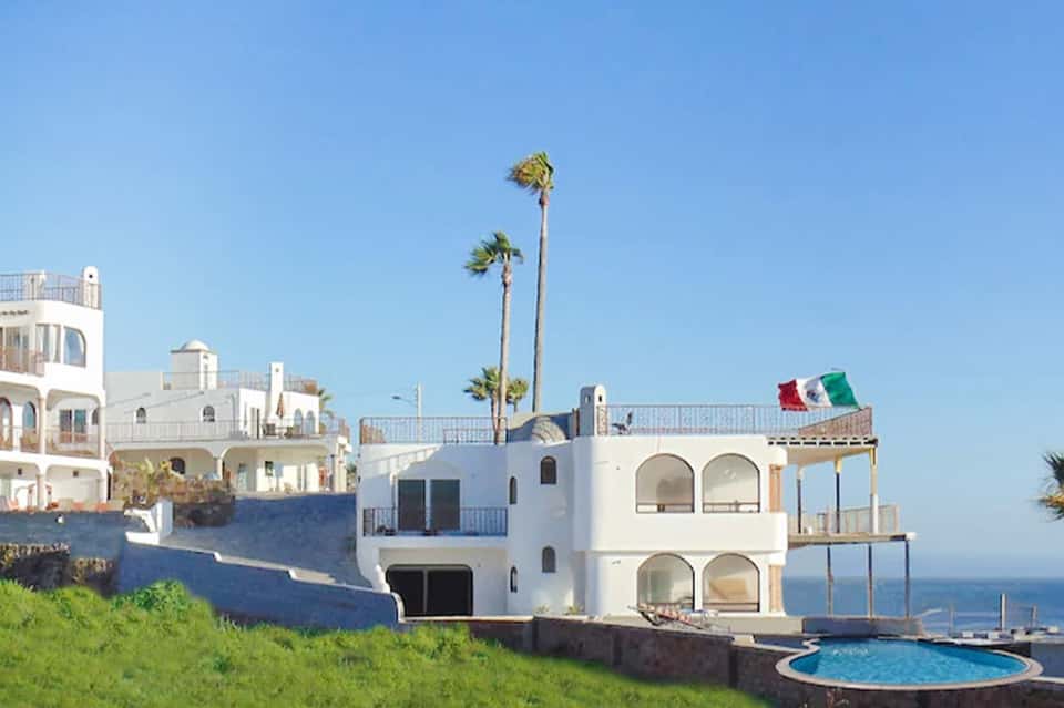 Clifftop white resort buildings with Mexican flag, palm trees, and circular pool overlooking ocean