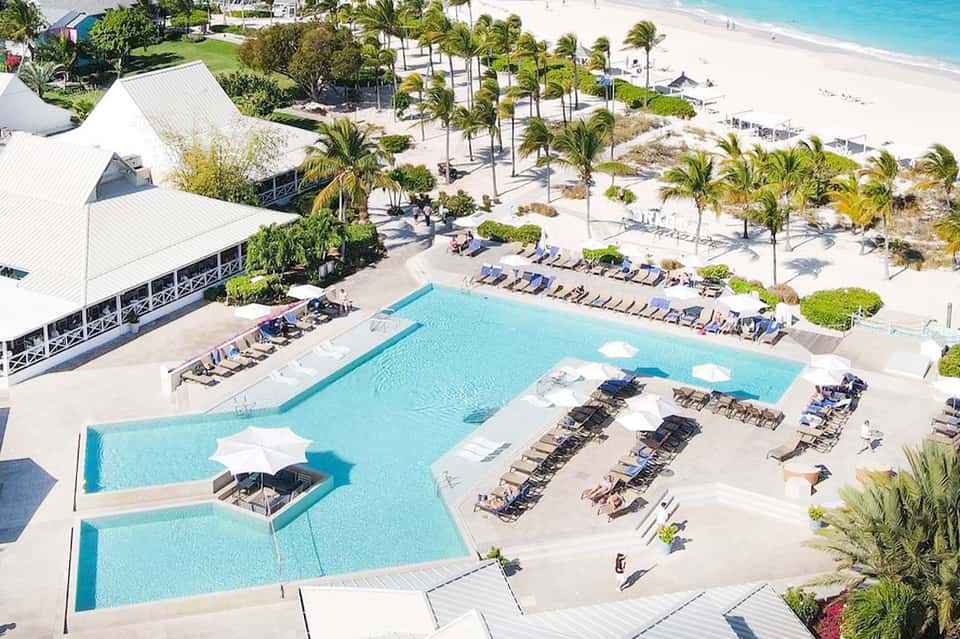 Aerial view of resort with turquoise pool, white structures, palm trees, and pristine beach