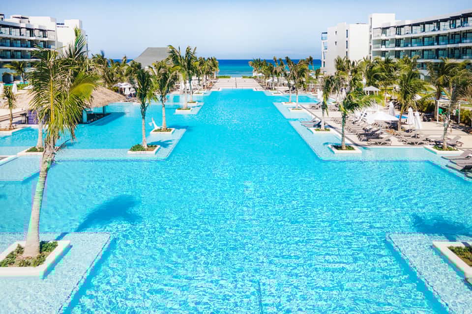 Aerial view of expansive turquoise resort pool with palm trees, lounge chairs, and ocean beyond
