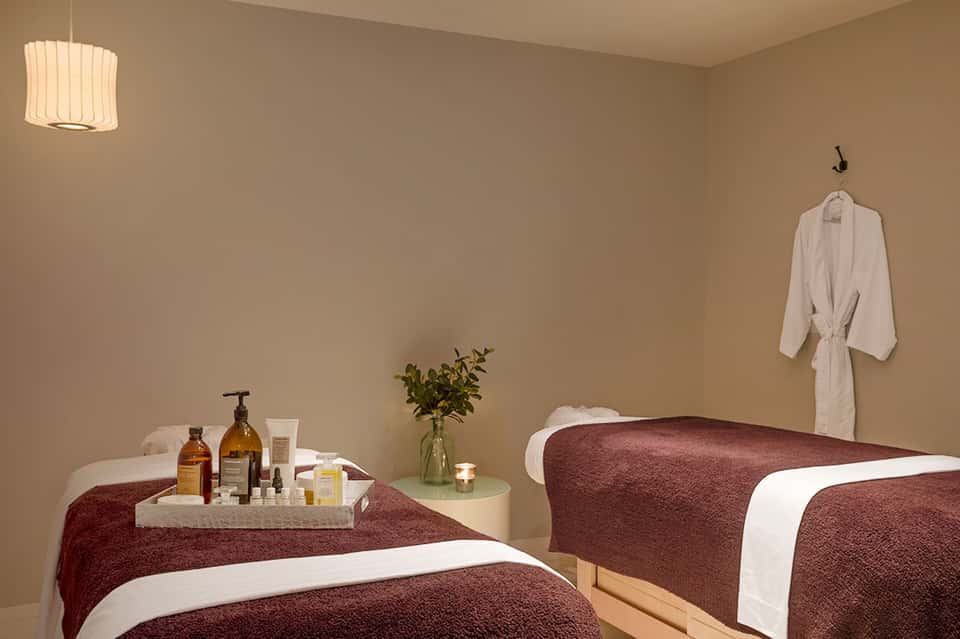 Spa treatment room with two massage beds, amber bottles, fresh flowers, and plush robes