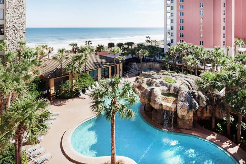 Curved resort pool with rock features and waterfall overlooking beach and pink hotel building