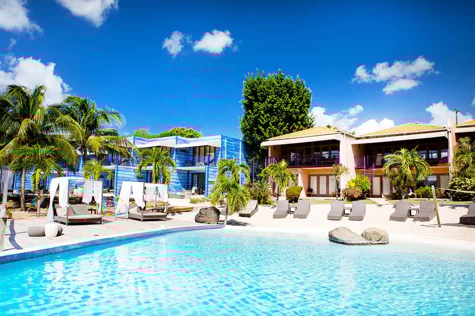 Resort pool with colorful blue and white buildings, palm trees, and lounge chairs under sunny sky