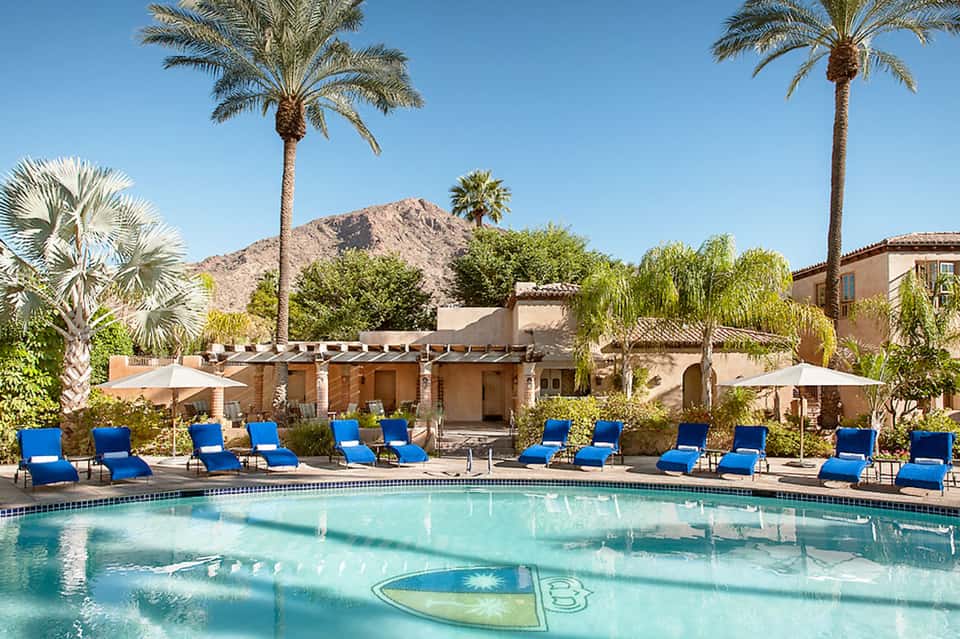 Desert resort pool with blue loungers, mountain backdrop, palm trees, and poolside cabana