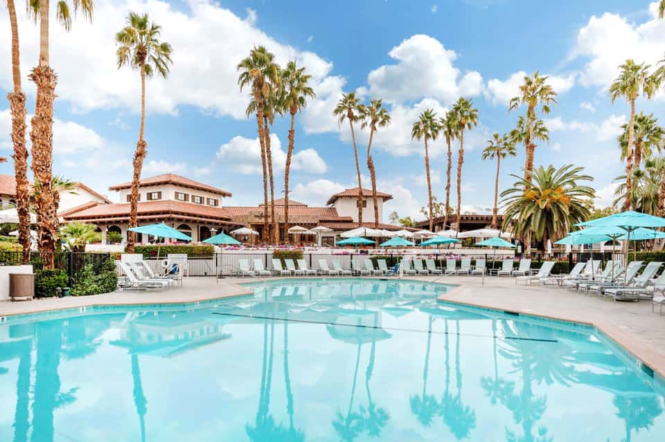 Resort pool with turquoise umbrellas, palm trees, Spanish colonial architecture, and white lounge chairs
