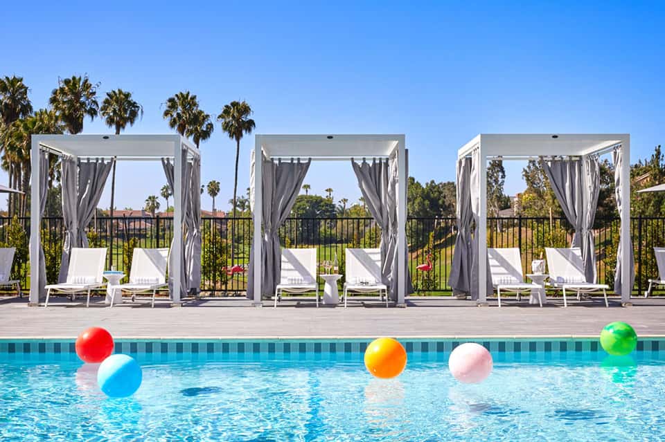 Resort pool with colorful inflatable balls and white cabanas with gray curtains overlooking palm trees
