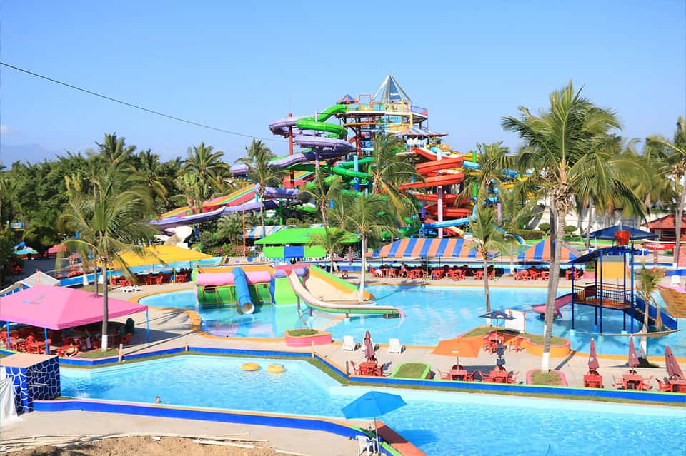 Colorful water park with tall slide structures, pools, and palm trees amid bright umbrellas