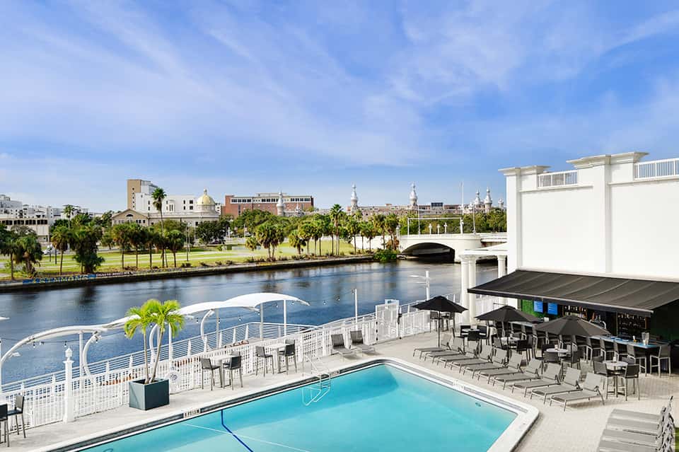 Waterfront resort pool with retractable roof bar, lounge chairs, and city skyline view