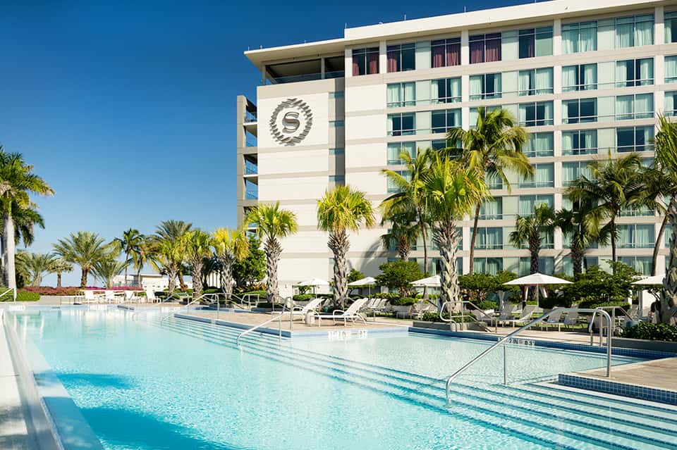 Resort lap pool with clear turquoise water, loungers, palm trees, and cream-colored hotel building