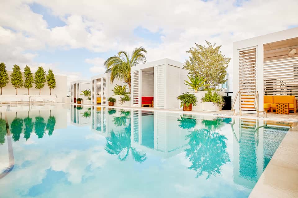 Modern resort infinity pool with white buildings, colorful lounge chairs, tropical landscaping, and reflective water surface