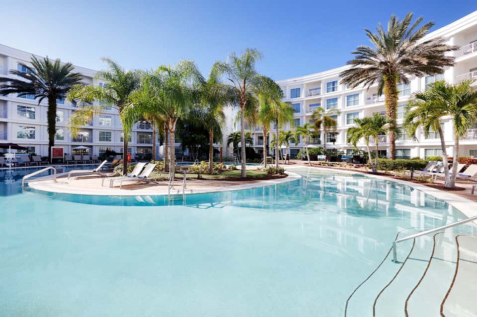 Resort pool surrounded by palm trees, white hotel buildings, and lounge chairs