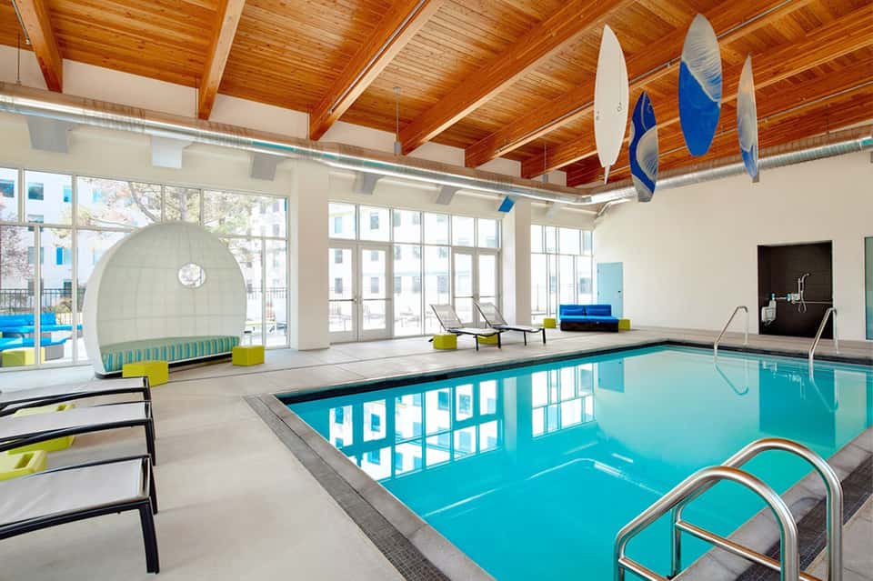 Indoor lap pool with wooden ceiling, large windows, modern seating, and blue surfboard decor