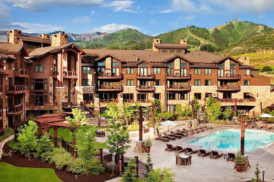Mountain resort with rustic lodge buildings, outdoor pool, lounge chairs, and green landscaping surrounded by forested peaks