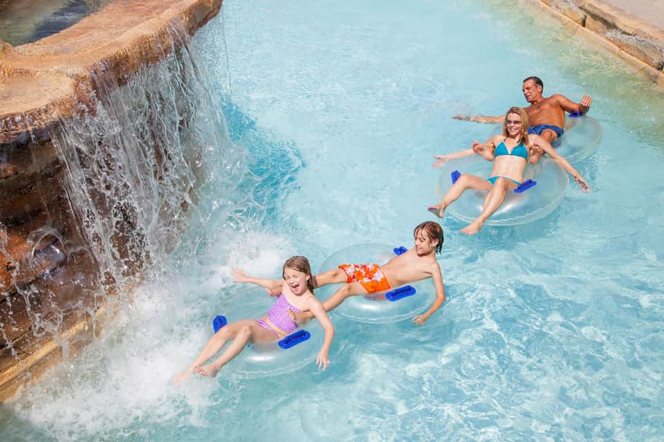 Family enjoying water slide and floats in bright turquoise resort pool with waterfall feature