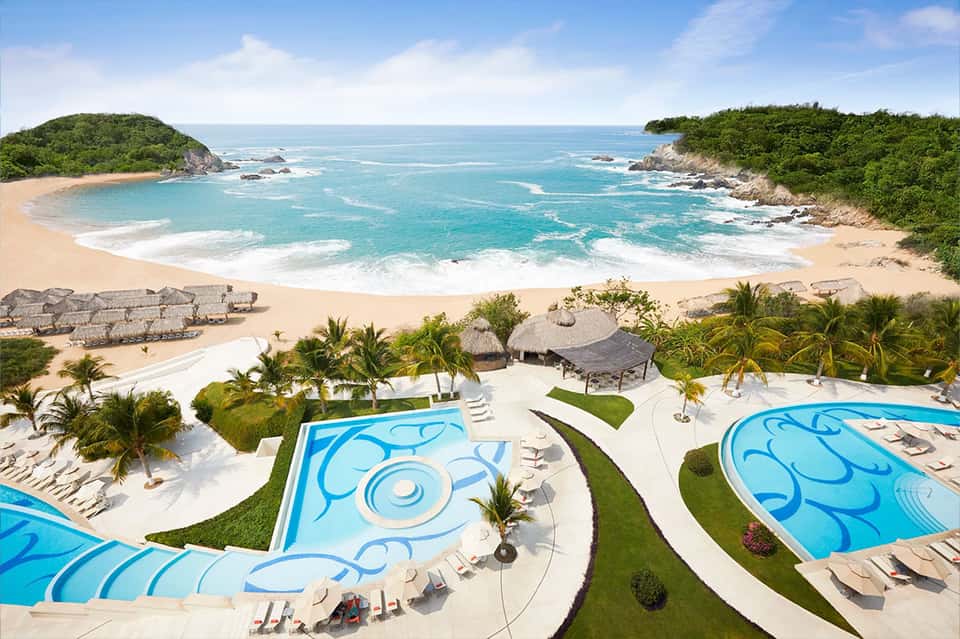Aerial view of curved resort pools overlooking a secluded cove beach with turquoise waters