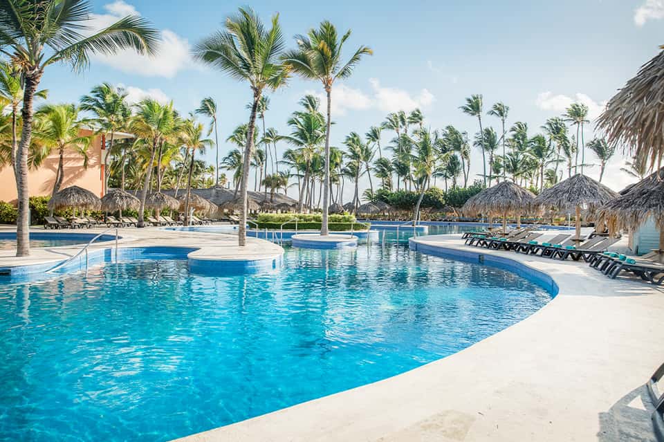 Resort pool complex with palm trees, tiki umbrellas, lounge chairs, and white sandy beach area