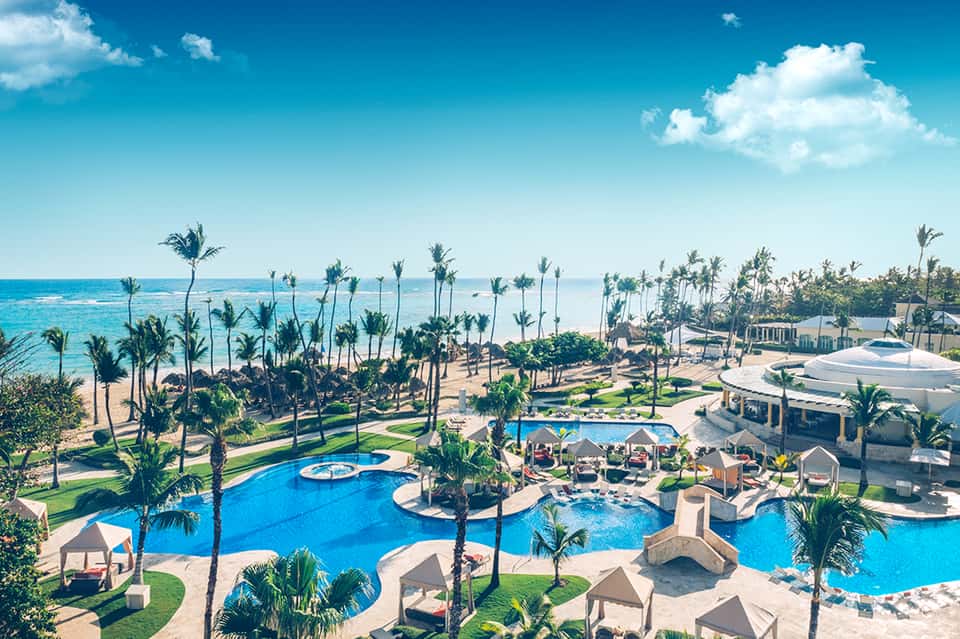 Aerial view of tropical beachfront resort with multiple blue pools, palm trees, and white buildings