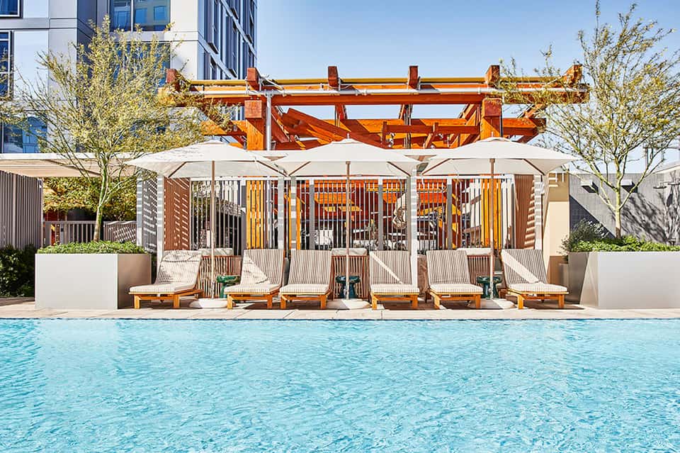Resort pool with white umbrellas and cushioned lounge chairs beneath wooden pergolas and modern high-rise buildings