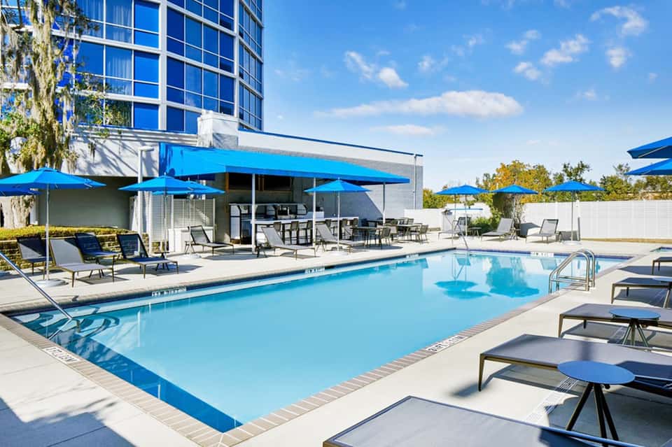 Modern hotel pool with blue umbrellas, lounge seating, glass high-rise building, and patio area