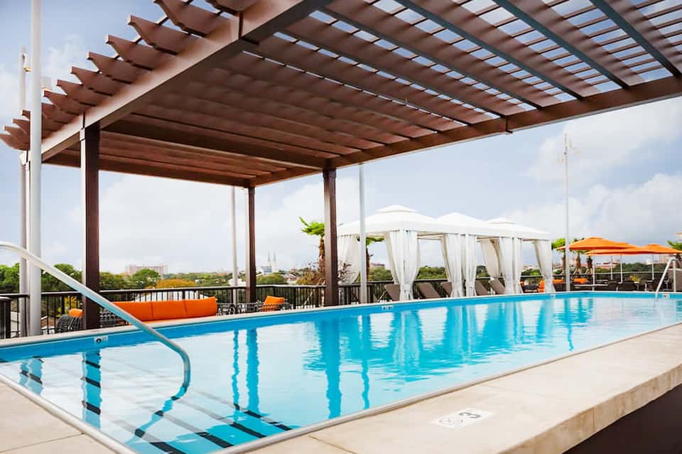 Rooftop infinity pool with pergola, loungers, white umbrellas, and distant cityscape