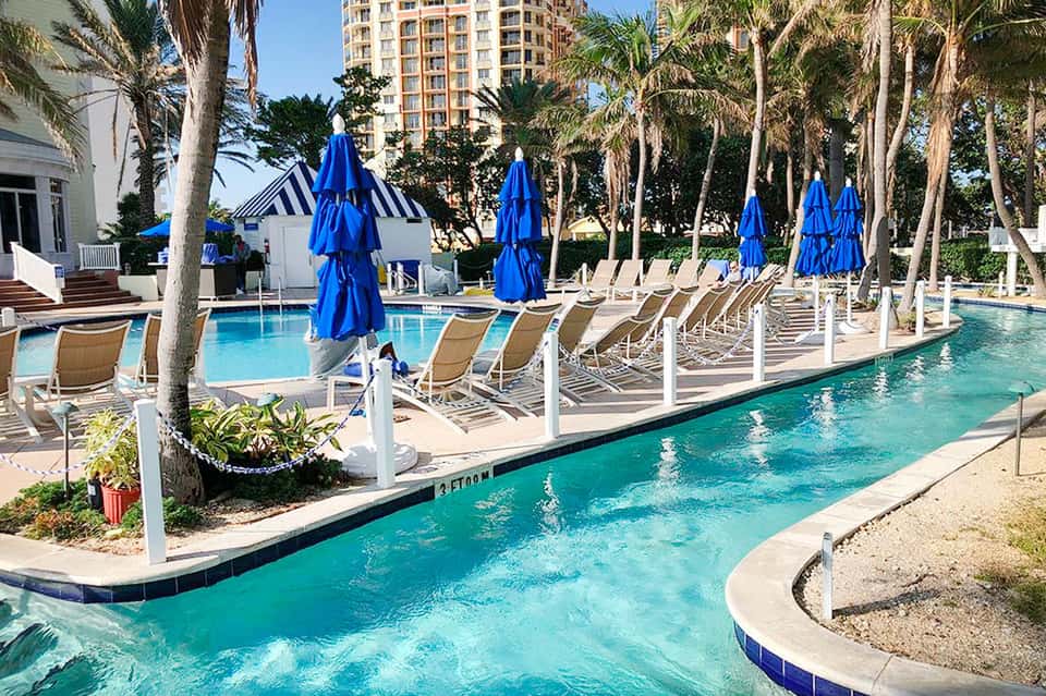 Resort lazy river with blue umbrellas, lounge chairs, and palm trees surrounding the water feature